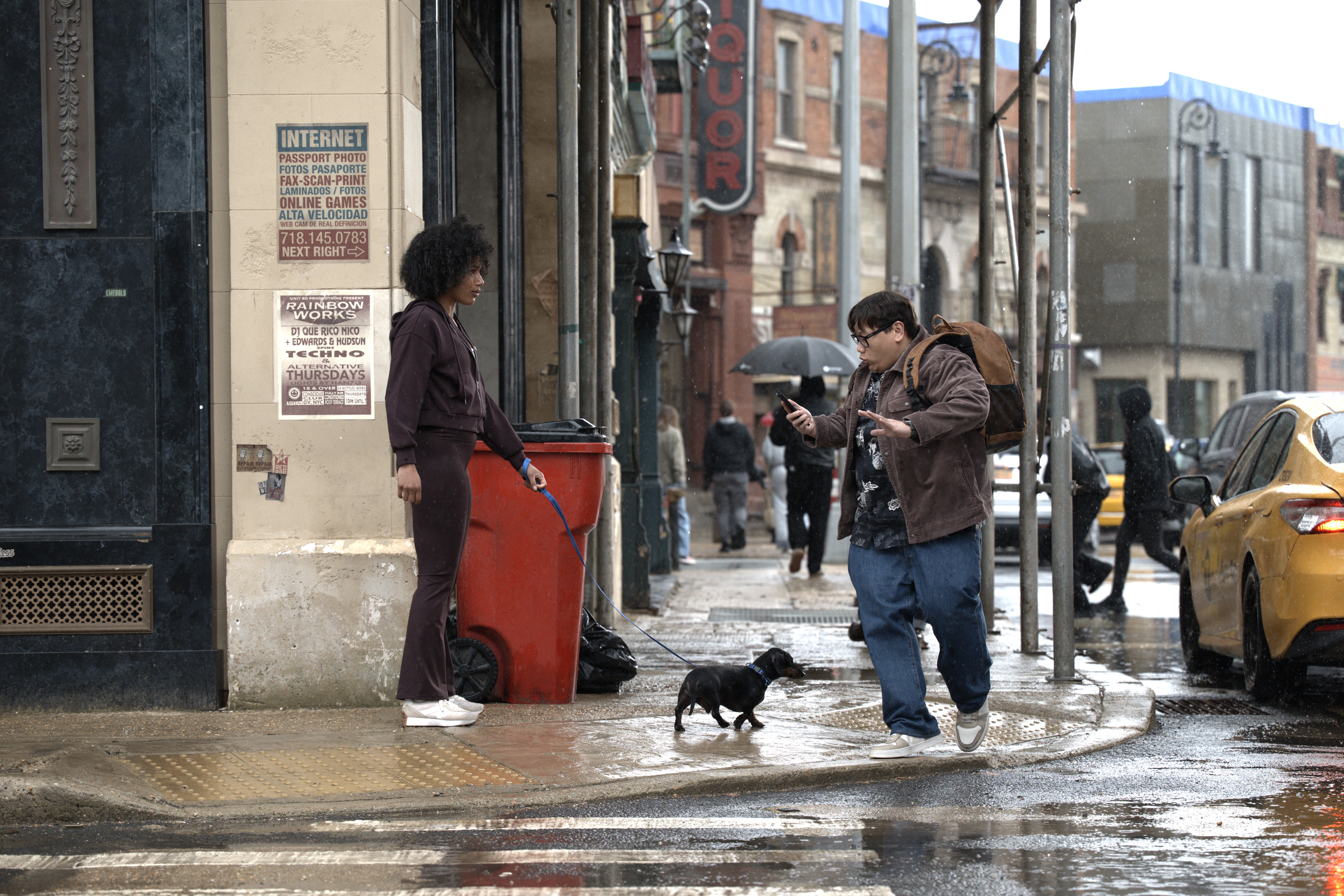 A person standing next to a red trash can on a rainy city street, with a small dog walking nearby and a person using a mobile device in the background.
