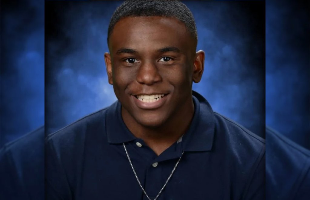 A smiling young Black man wearing a navy blue shirt against a blue background.