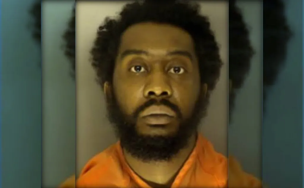 A close-up portrait of a Black man with a beard wearing an orange prison jumpsuit, looking directly at the camera with a serious expression.