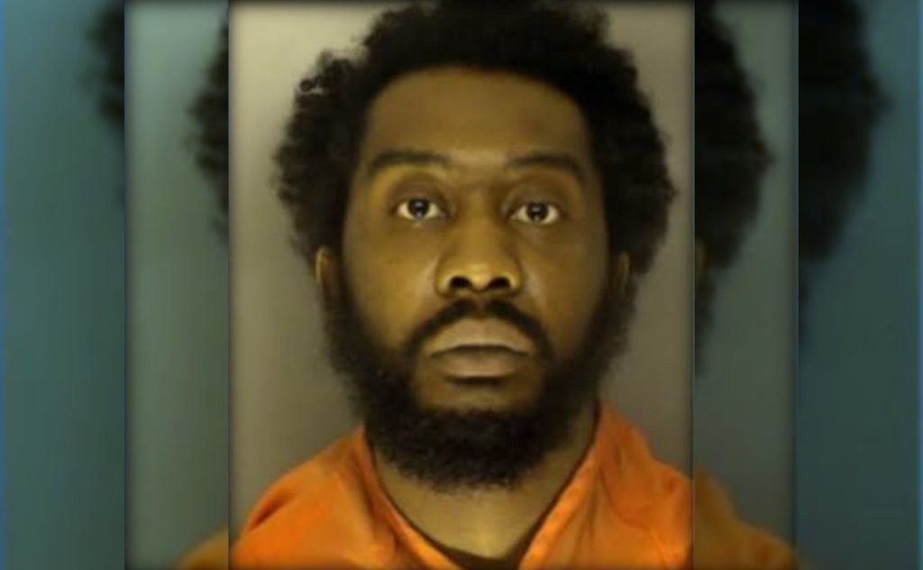 A close-up portrait of a Black man with a beard wearing an orange prison jumpsuit, looking directly at the camera with a serious expression.