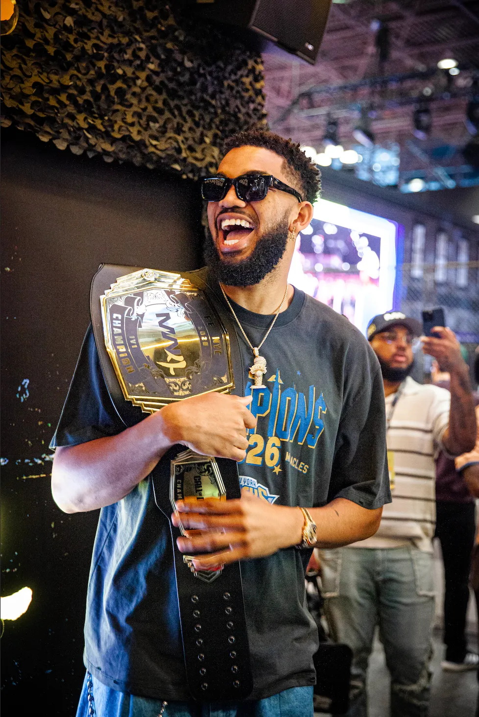 A man with a curly beard and sunglasses laughing while holding a championship belt with "MVP" written on it, surrounded by people taking photos.