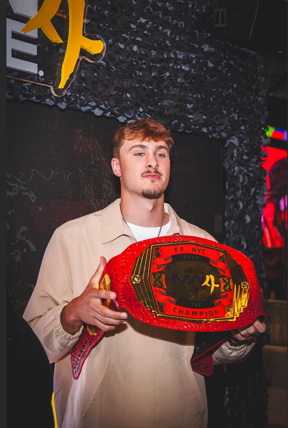 A young man wearing a beige jacket holds a red championship wrestling belt with the text "FF NYC CHAMPION" in front of a dark background with yellow and red accents.