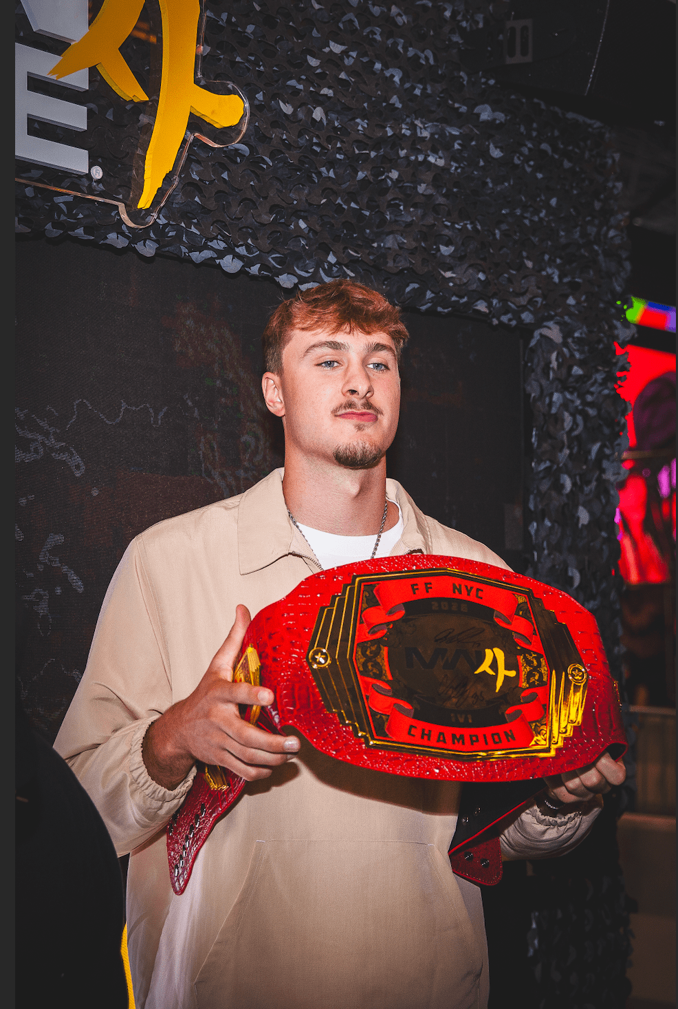 A young man wearing a beige jacket holds a red championship wrestling belt with the text "FF NYC CHAMPION" in front of a dark background with yellow and red accents.