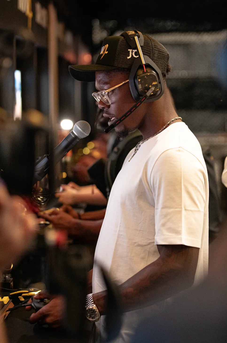 A person wearing a black cap with "JC" logo, headphones, and a white shirt speaking into a microphone on a stage.