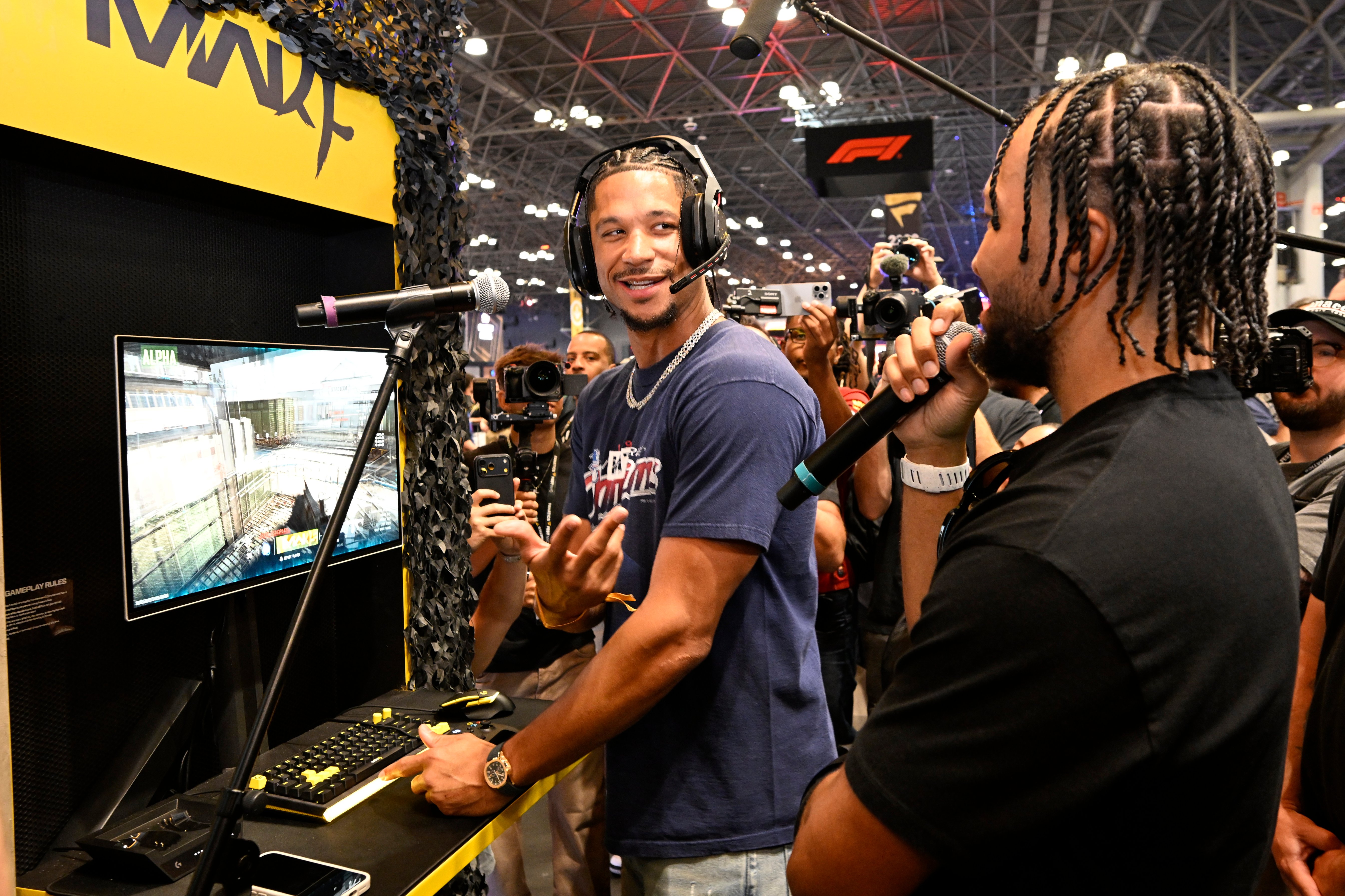 Two men, one wearing a headset, playing a video game on a large screen at a gaming event.