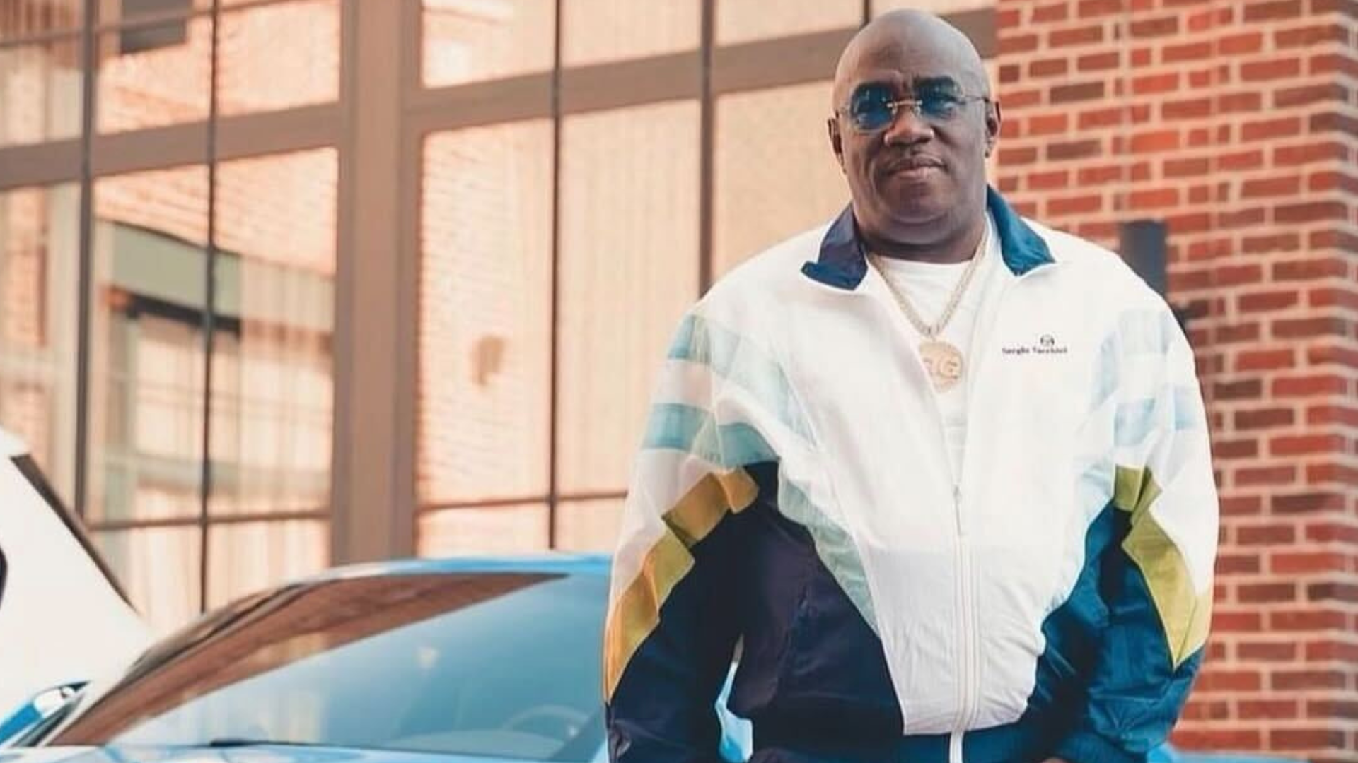 A middle-aged man wearing a white jacket with blue and yellow stripes, standing in front of a brick building and a blue car.
