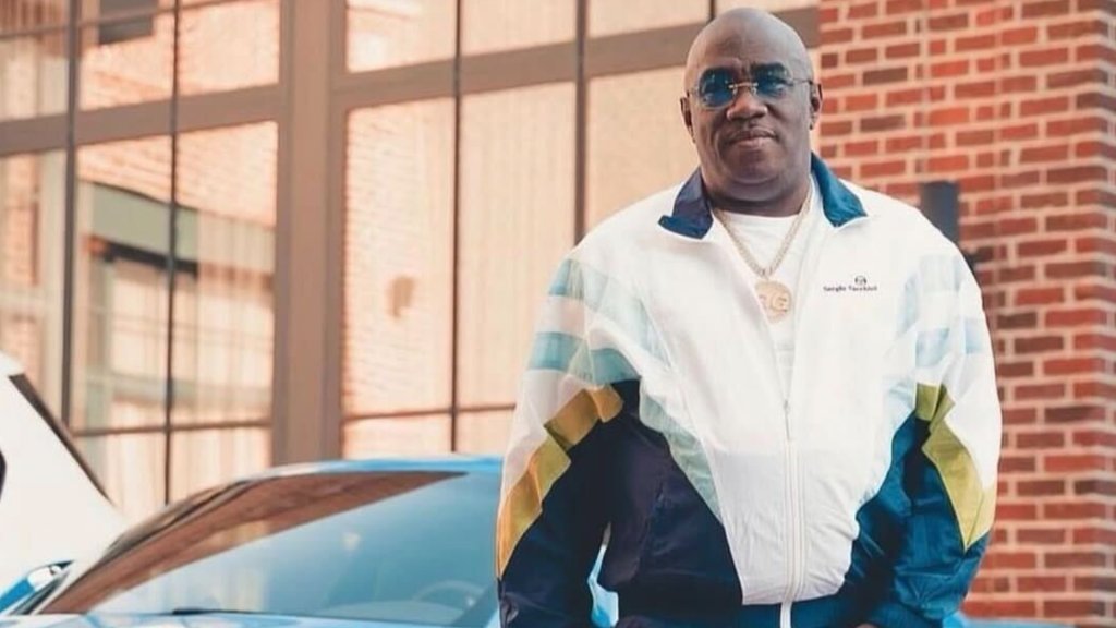 A middle-aged man wearing a white jacket with blue and yellow stripes, standing in front of a brick building and a blue car.