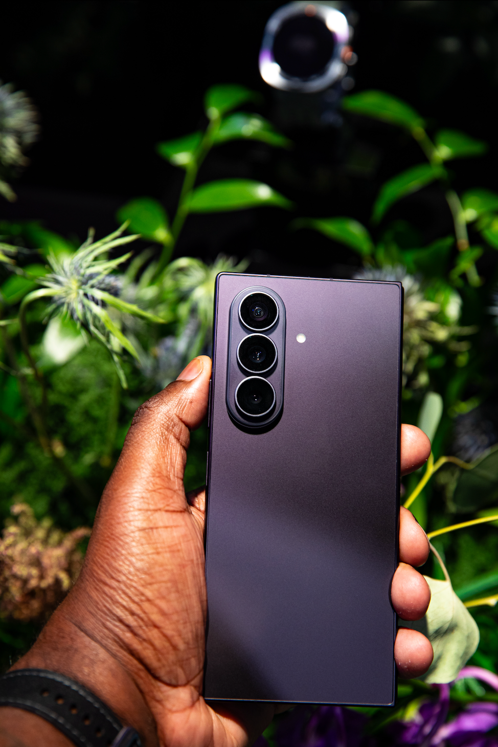 A hand holding a black smartphone with a triple camera lens against a backdrop of lush green foliage.