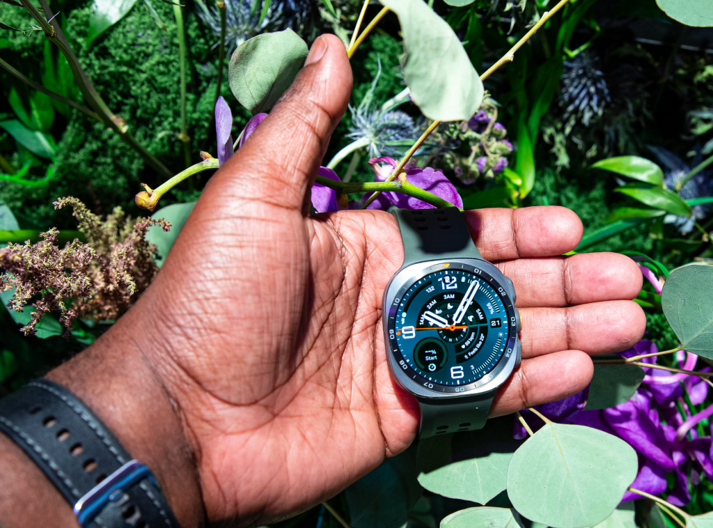 A person's hand holding a black and blue analog watch against a background of lush green foliage and purple flowers.