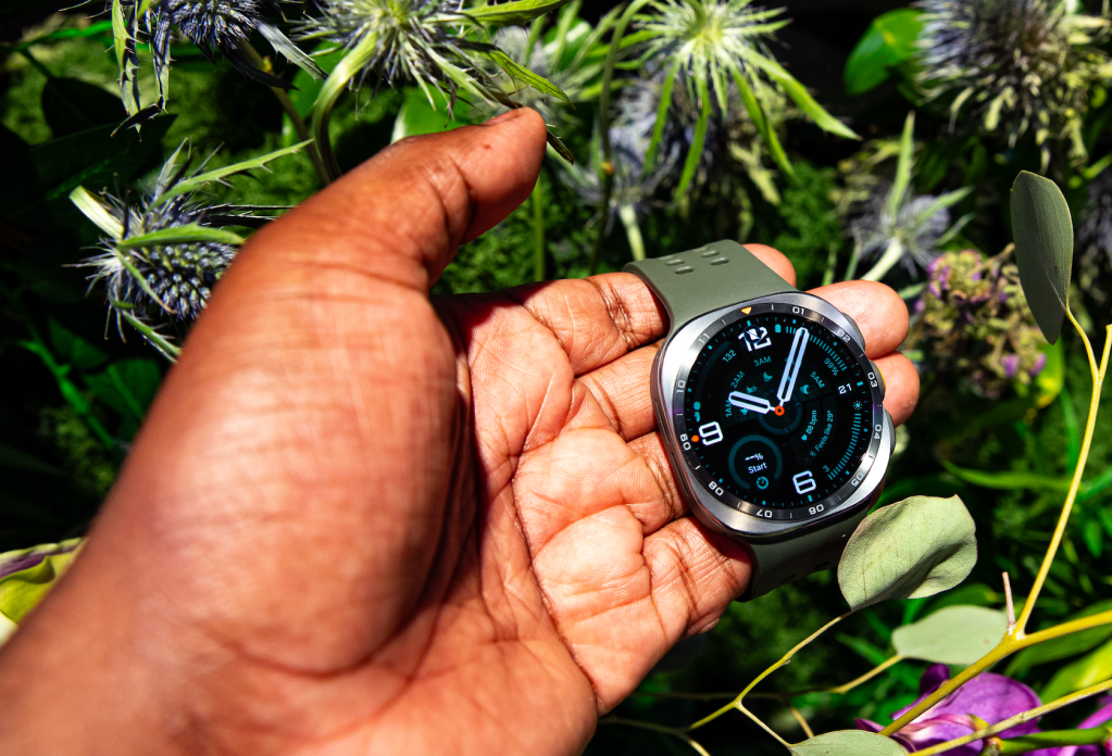 A person's hand holding a smartwatch displaying the time in a lush, green outdoor setting with purple flowers.