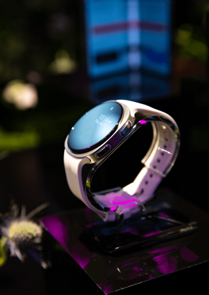 A close-up view of a smartwatch with a round face and a purple band, resting on a dark surface with blurred greenery in the background.