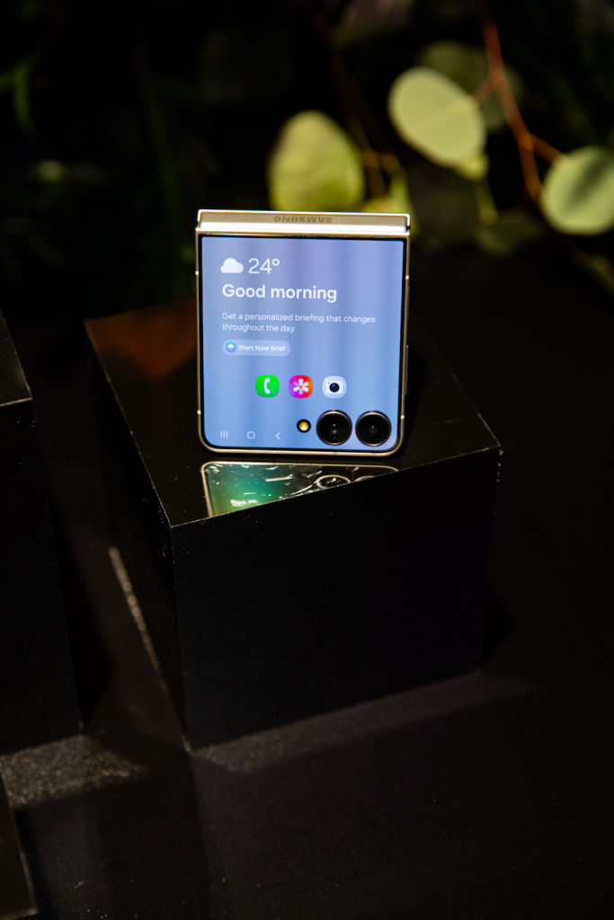 A smartphone displaying the time and weather information on a dark background with blurred greenery in the background.