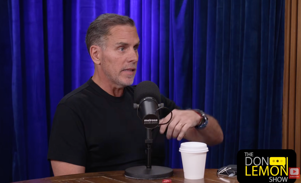 A man with gray hair speaking into a microphone on a stage with a blue curtain backdrop, wearing a black t-shirt with the Melrose Studios logo.