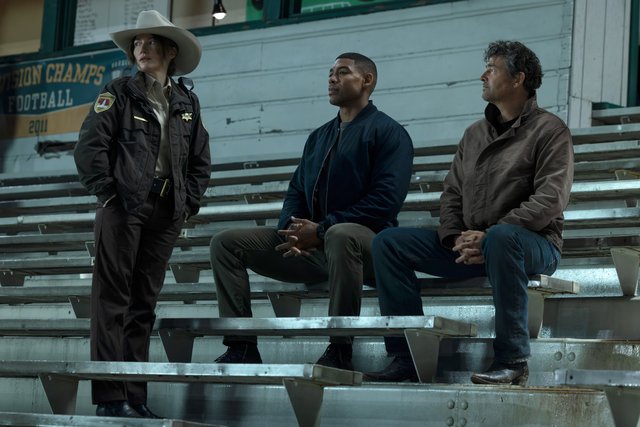 Three men sitting on stadium bleachers, one wearing a cowboy hat.