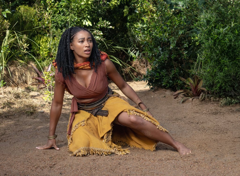 A person wearing a brown top and yellow skirt sits on a dirt path surrounded by lush greenery.