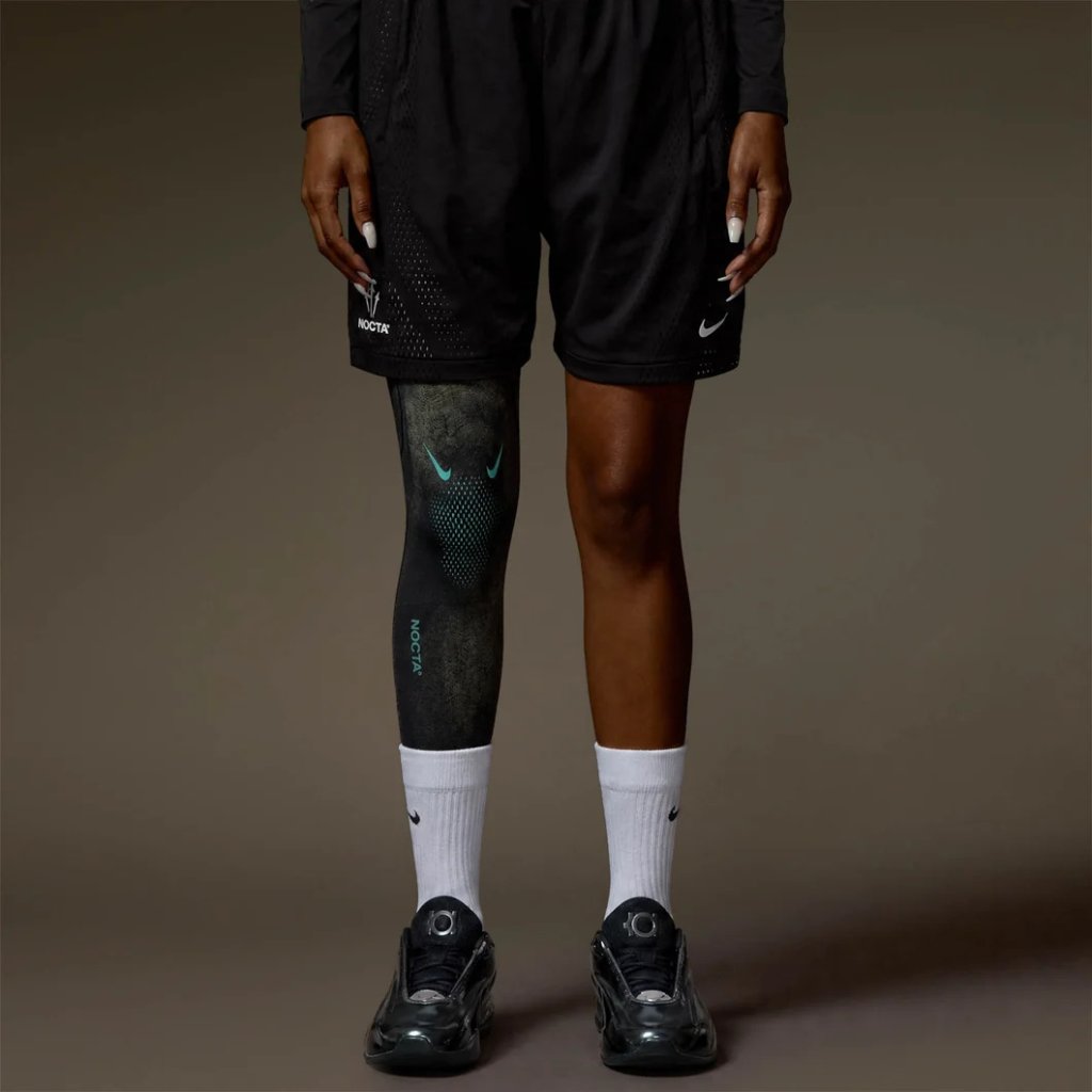 A person wearing black Nike athletic shorts and socks, with a Nike Nocta logo on the leg, standing against a dark background.