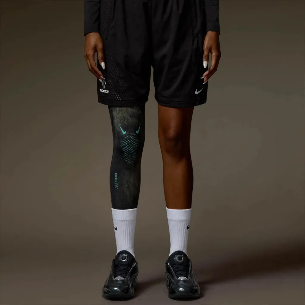 A person wearing black Nike athletic shorts and socks, with a Nike Nocta logo on the leg, standing against a dark background.