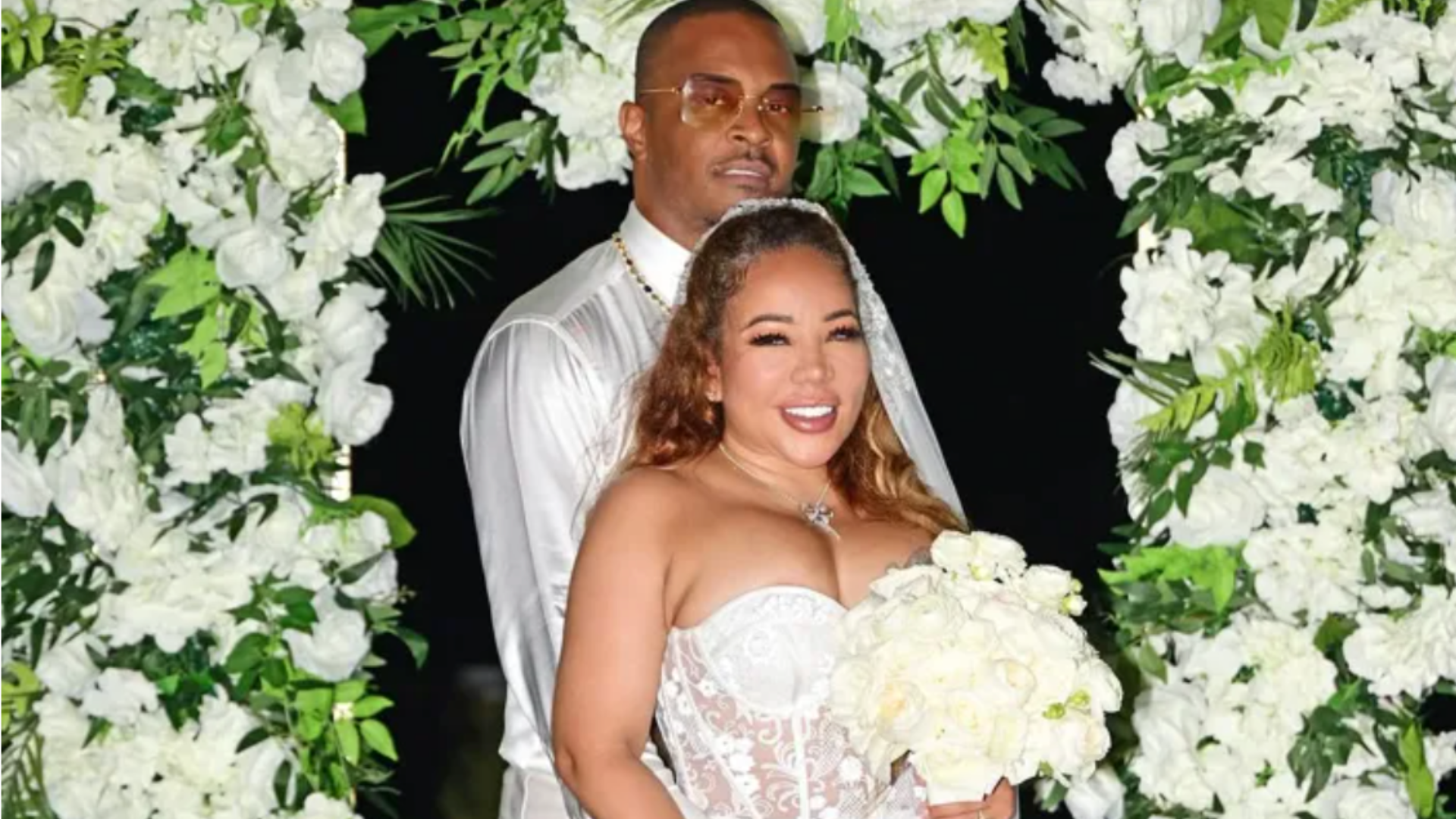 A bride and groom standing in a floral setting, the bride holding a bouquet of white flowers and smiling.