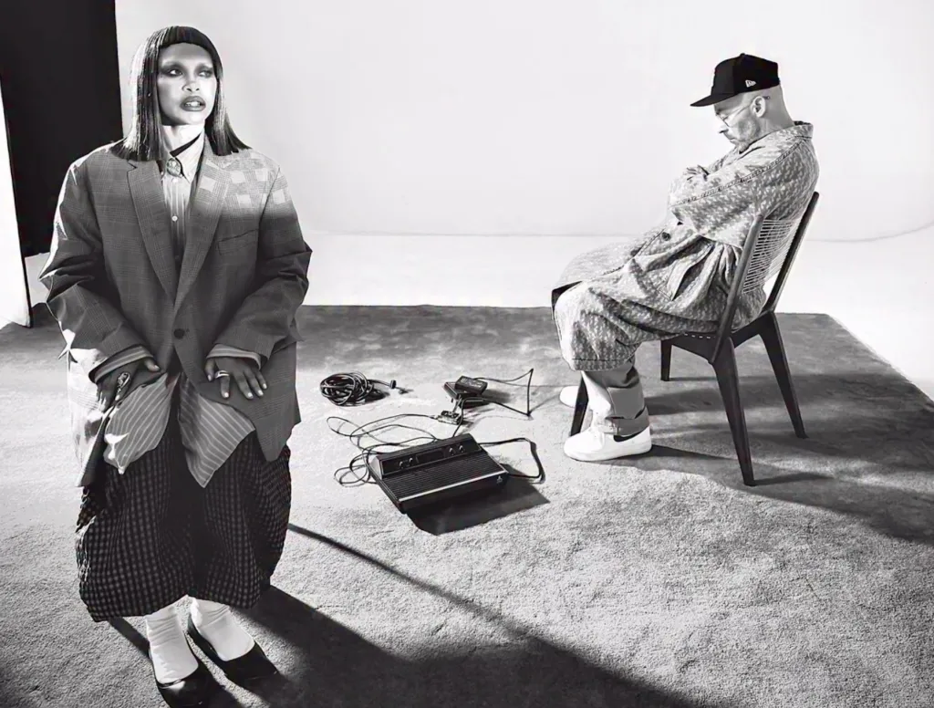 Two people in a studio setting, one standing and one seated, with various audio equipment on the floor between them.