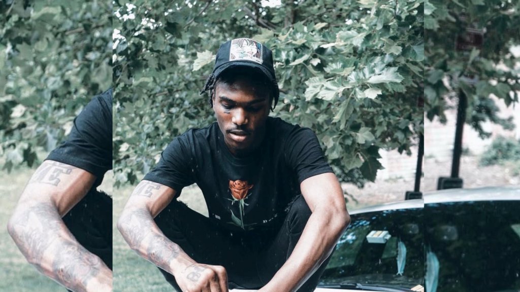 A person wearing a black cap and shirt, sitting on the ground next to a vehicle in a wooded area.