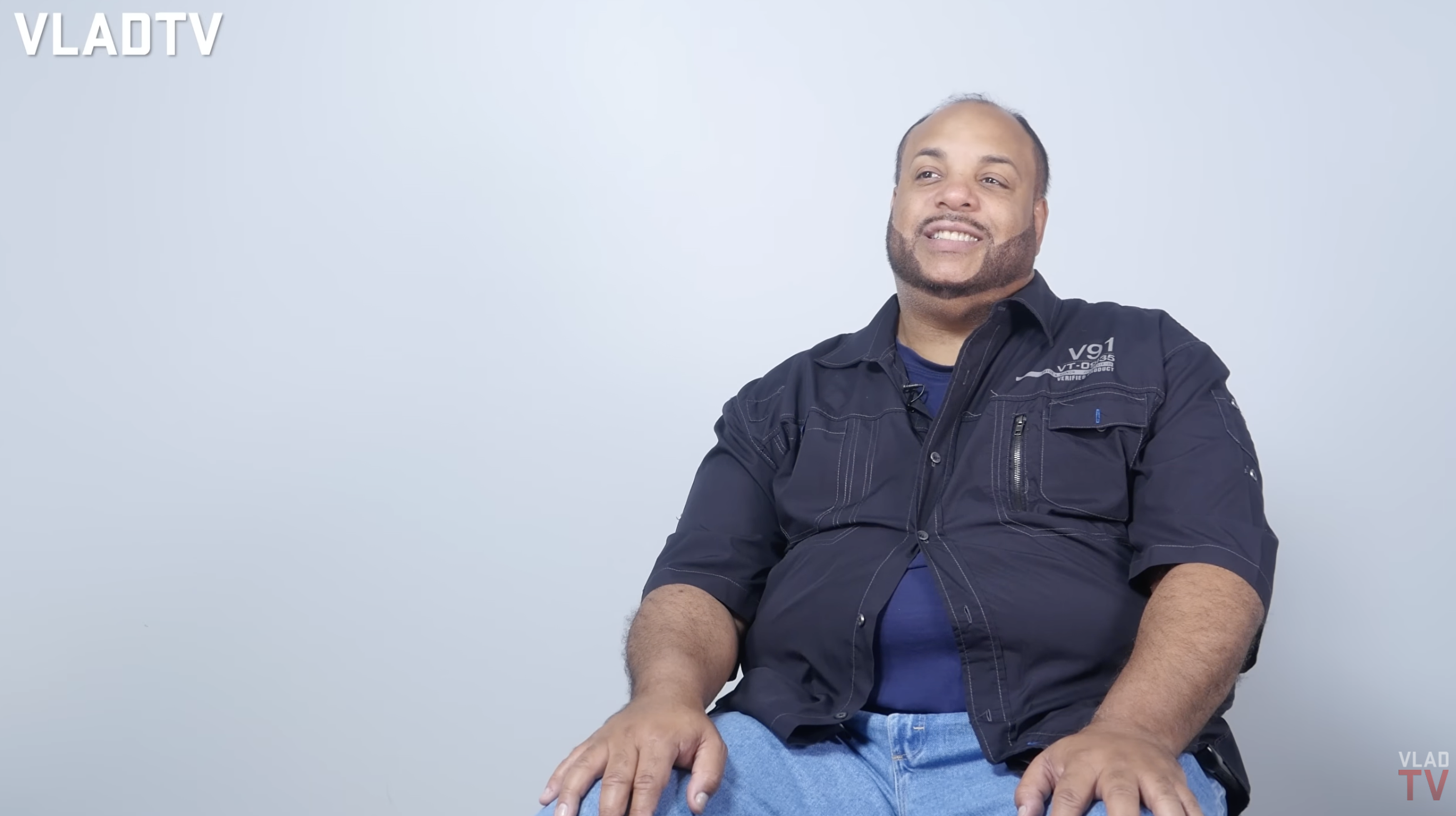 A smiling man wearing a black jacket with pockets and a zipper, sitting against a plain background.