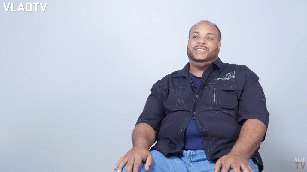 A smiling man wearing a black jacket with pockets and a zipper, sitting against a plain background.