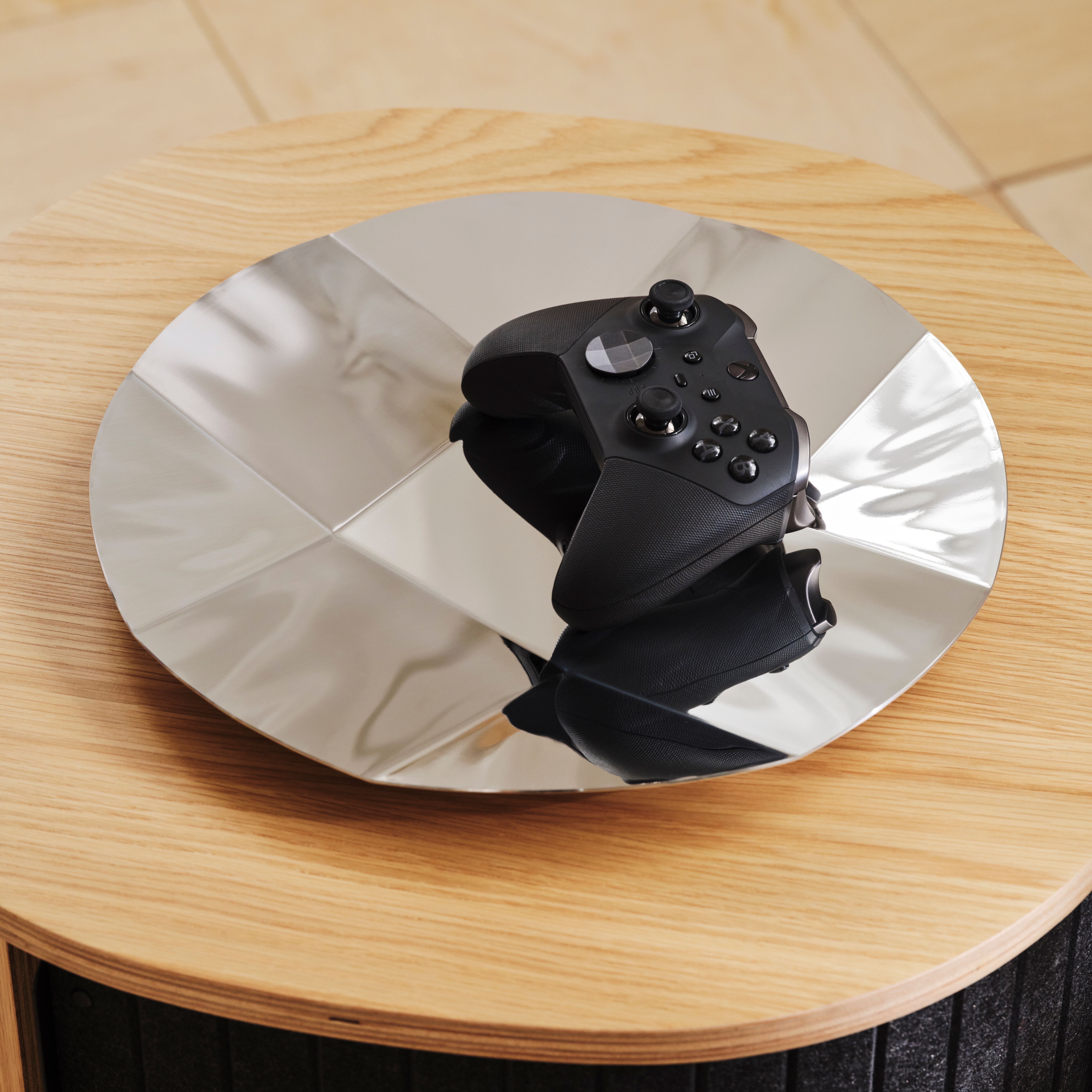 A black video game controller resting on a reflective silver surface on a wooden table.