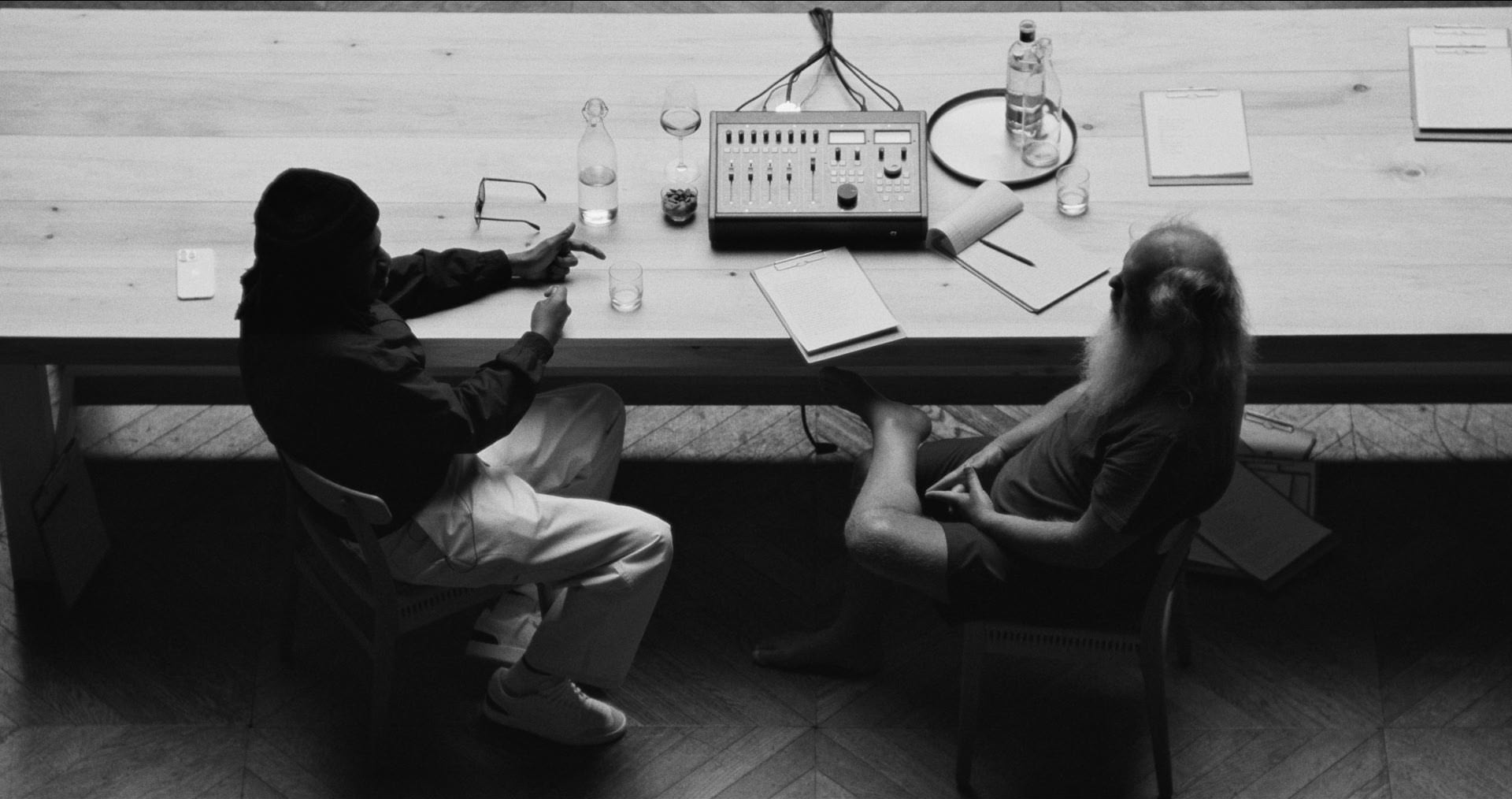 Two people sitting at a desk with various audio equipment and papers.